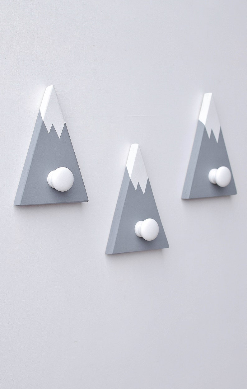 May include: Three gray wooden mountain-shaped hooks with white snow caps and white knobs. The hooks are mounted on a white wall.