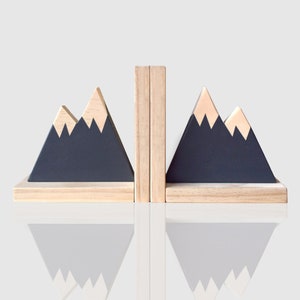 Puede incluir: Un par de sujetalibros de madera con forma de montañas con detalles pintados en negro y beige. Los sujetalibros están hechos de madera natural y tienen un aspecto rústico.