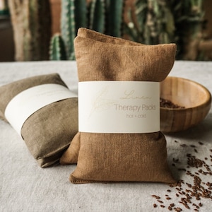 Könnte beinhalten: Zwei braune Leinen-Therapiepackungen mit weißen Etiketten mit der Aufschrift "Linen Therapy Packs hot + cold". Eine Packung ist hellbraun, die andere dunkelbraun. Eine Holzschale und Samen sind im Hintergrund.