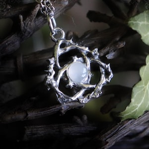 Op de afbeelding: Zilverkleurige hangerketting met een lichtgekleurde, ronde steen in het midden, gezet in een gedetailleerd, doornig, takachtig ontwerp. De ketting heeft een fijne ketting. Het Witching Hour-logo staat onderaan.
