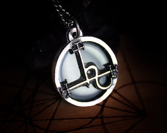 May include: A silver pendant necklace with a circular design featuring a black symbol inside. The symbol is a stylized letter "L" with a circle inside the letter. The pendant is on a black chain.