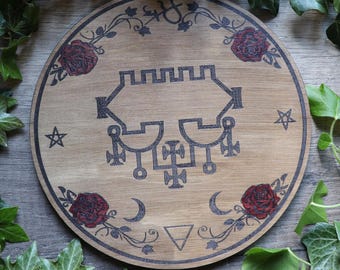 May include: A round wooden disc with a black and white sigil in the center. The sigil is surrounded by a border of roses and vines. The disc is on a wooden surface.