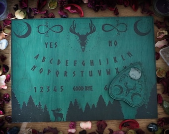 Könnte beinhalten: Ein grünes Ouija-Brett aus Holz mit einem Planchette, das einen Hirschschädel und Mondsicheln zeigt. Das Brett ist mit dem Alphabet, den Zahlen 1-6, "ja", "nein" und "auf Wiedersehen" bedruckt.