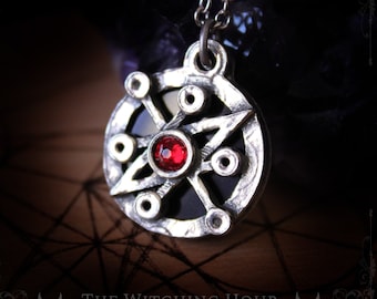 Puede incluir: Collar con colgante de plata con una piedra preciosa roja en el centro de un diseño de estrella. El colgante está rodeado por un círculo de círculos más pequeños.