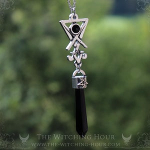 May include: A silver necklace with a black gemstone pendant. The pendant features a pentagram and a sigil. The necklace is against a green background.