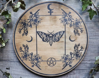 May include: A wooden circle with a black and white design of a moth, flowers, a crescent moon, a pentagram, and the symbol for Venus. The circle is on a woodgrain background.