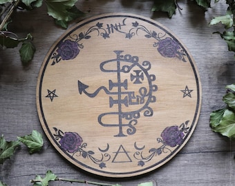 May include: A wooden circle with a black and white sigil in the centre. The sigil is surrounded by a floral design with purple roses. The circle is on a wooden background.