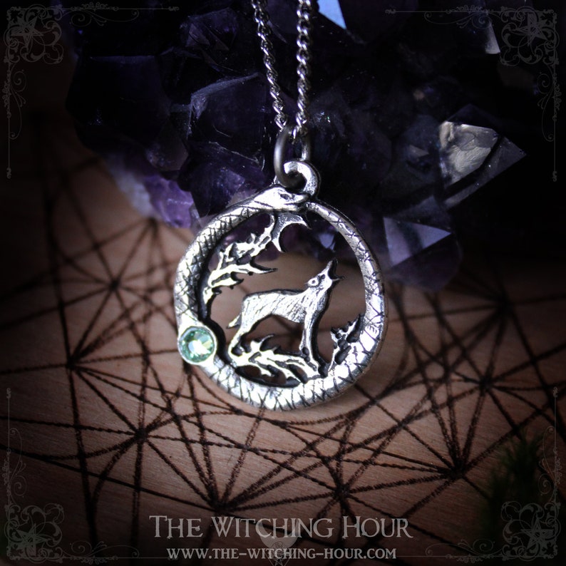 Puede incluir: Collar de plata con un colgante que representa un lobo aullando a la luna, rodeado de una serpiente y follaje. El colgante tiene un acento de piedra preciosa verde.