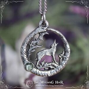 Puede incluir: Collar con colgante de plata que representa un lobo aullando a la luna, rodeado de una luna creciente y follaje. El colgante tiene un acento de piedra preciosa verde.