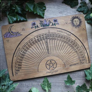 May include: A rectangular wooden dowsing board featuring a semi-circular scale with numerical values from 0 to 18,000. The board includes illustrations of a crescent moon, purple flowers, crystals, and a sun, with a central pentagram symbol.