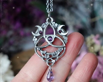 May include: A silver-toned metal pendant necklace featuring a central triquetra knot design, two crescent moons with small dark red stones, and a central purple stone. The pendant is adorned with leaf motifs and a dangling purple crystal drop.