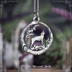 Puede incluir: Collar con colgante de plata que representa un lobo aullando a la luna, rodeado de una serpiente y follaje. El colgante tiene un acento de piedra preciosa verde.