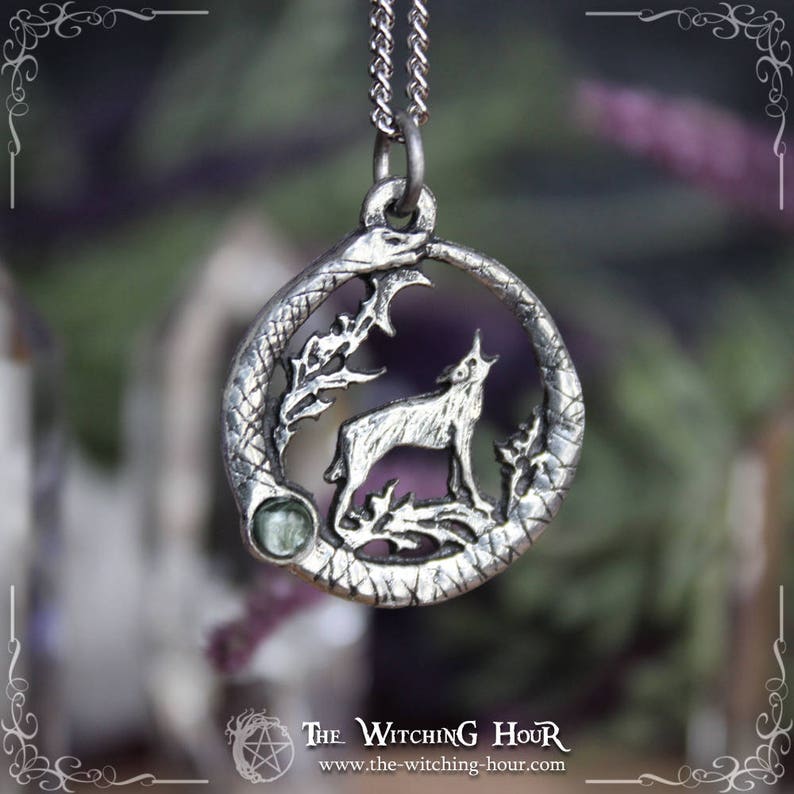 Puede incluir: Collar con colgante de plata que representa un lobo aullando a la luna, rodeado de una serpiente y follaje. El colgante tiene un acento de piedra preciosa verde.