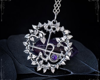 May include: A silver-toned circular pendant featuring an intricate vine and leaf border surrounding a central symbol and a small, round, purple gemstone. The pendant hangs from a silver-toned chain and is positioned over a dark purple crystal.