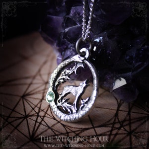 Puede incluir: Collar con colgante de plata que presenta un lobo aullando a la luna, rodeado de una serpiente, con un acento de piedra preciosa verde. El colgante est&aacute; en una cadena de plata y descansa sobre una superficie de madera con un patr&oacute;n geom&eacute;trico.