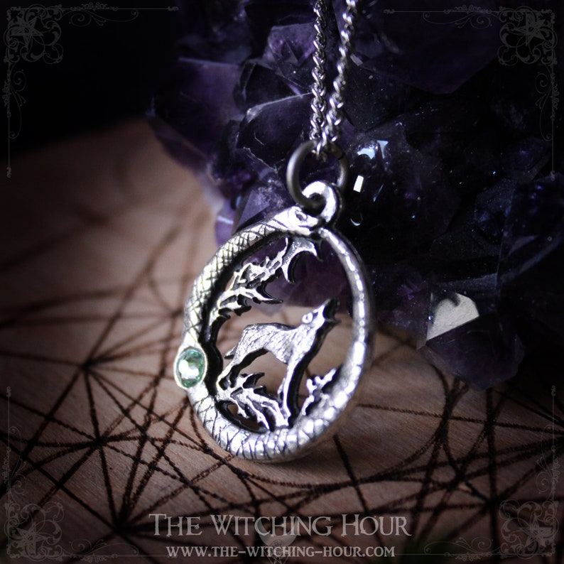Puede incluir: Collar con colgante de plata que presenta un lobo aullando a la luna, rodeado de una serpiente, con un acento de piedra preciosa verde. El colgante est&aacute; en una cadena de plata y descansa sobre una superficie de madera con un patr&oacute;n geom&eacute;trico.