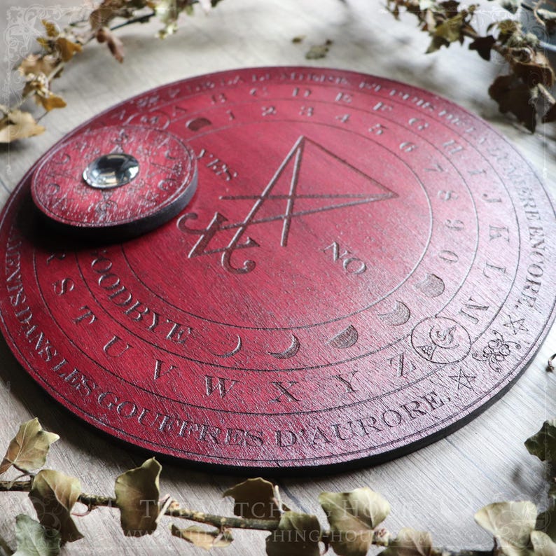 May include: A red wooden circular divination board with a black triangle and a smaller circular piece with a crystal in the center. The board has a circle of numbers, letters, and moon phases around the edge.