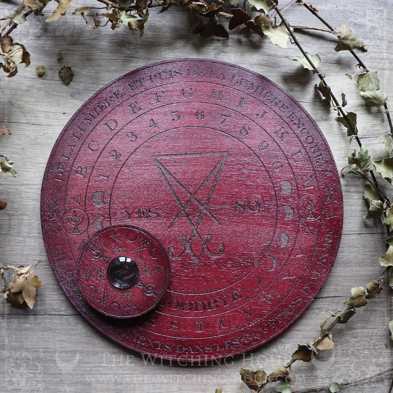 May include: A red wooden Ouija board with a black pentagram and the words "DE LA LUMIERE ETUIS DE LA LEMIERE ENCORE" around the edge. The board has a smaller, circular planchette with a glass window.