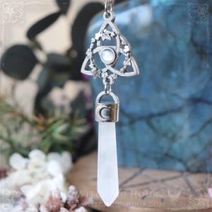 Può includere: Collana con pendente argentato con un motivo a nodo celtico triangolare, dettagli floreali e una pietra di luna centrale. Sospende una punta di cristallo di quarzo trasparente. La collana ha una catena d'argento e una chiusura con una luna crescente.