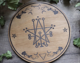 Può includere: Un piatto rotondo in legno con un design a pentagramma in bianco e nero. Il piatto è circondato da un bordo decorativo di rose, lune e stelle. Il testo "The Witching Hour" è stampato nella parte inferiore del piatto.