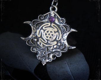 May include: A silver-toned pendant on a chain featuring an intricate embossed design with a central circular motif, a small five-pointed star, and a single purple gemstone at the top. The text THE WITCHING HOUR and WWW.THE-WITCHING-HOUR.COM is visible.