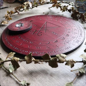 May include: A red wooden Ouija board with a black planchette and a pentagram design. The board has a black border and is surrounded by dried leaves.
