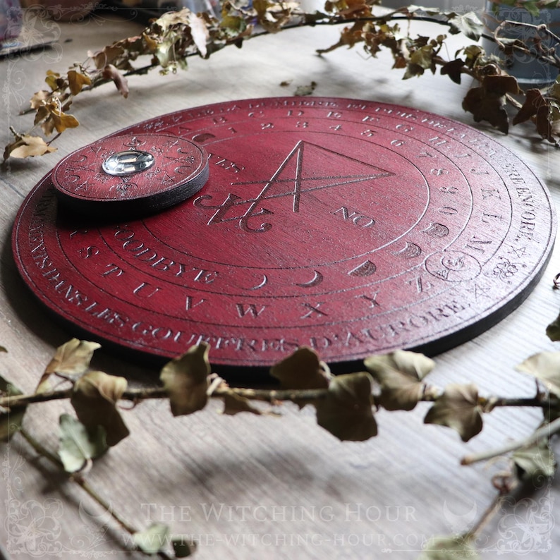 May include: A red wooden Ouija board with a black planchette and a pentagram design. The board has a black border and is surrounded by dried leaves.