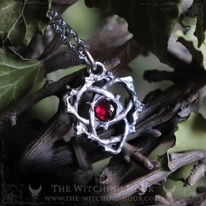 May include: A silver pendant necklace featuring a deep red gemstone. The pendant has an intricate, organic design. The chain is silver. The Witching Hour logo and website address are visible at the bottom.