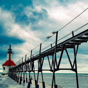 Puede incluir: Un faro rojo y blanco se alza sobre un largo muelle que se extiende hacia un mar azul. El cielo es una mezcla de azul y gris con nubes esponjosas. Gaviotas vuelan por encima.