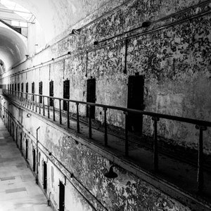 May include: A black and white photo of a long, narrow hallway in a prison. The hallway is lined with barred cell doors. The walls are cracked and peeling, and the floor is dirty. The photo is taken from a high angle, looking down the hallway.
