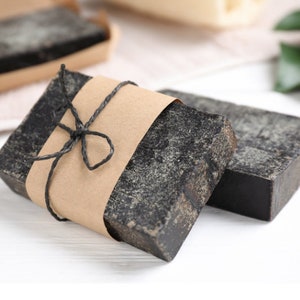 Può includere: Due saponette di carbone nero avvolte in carta marrone con un fiocco di spago nero.