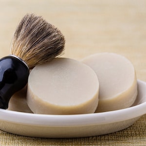 Puede incluir: Tres pastillas de jabón de afeitar redondas de color blanco roto en un plato de cerámica blanco con una brocha de afeitar negra y plateada.