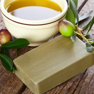 Puede incluir: Una barra de jabón de aceite de oliva verde junto a un tazón de aceite de oliva y una rama de olivo con aceitunas verdes.