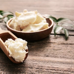 May include: A wooden bowl filled with white shea butter, with a wooden scoop containing more shea butter. The bowl and scoop are on a wooden surface.