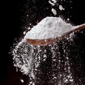 Op de afbeelding: Een houten lepel vol wit poeder, met enkele poederdeeltjes in de lucht. De achtergrond is effen zwart, wat het contrast en de textuur van het poeder benadrukt.