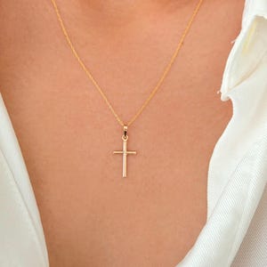 May include: A delicate gold chain necklace featuring a simple, polished gold cross pendant. The necklace rests against the neck, showcasing a thin, refined metal link chain that connects to a small, vertical cross charm with clean, straight edges.