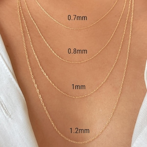 Könnte beinhalten: Nahaufnahme von vier goldenen Halsketten mit unterschiedlichen Kettendicken: 0,7 mm, 0,8 mm, 1 mm und 1,2 mm. Die Ketten werden vor einem weißen Hemd präsentiert, um die Größenunterschiede zu zeigen.