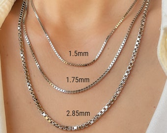 May include: Three silver-toned box chain necklaces of varying thicknesses, labeled with their respective widths of 1.5mm, 1.75mm, and 2.85mm. The chains feature a square link design and are worn layered, showcasing the difference in link size.
