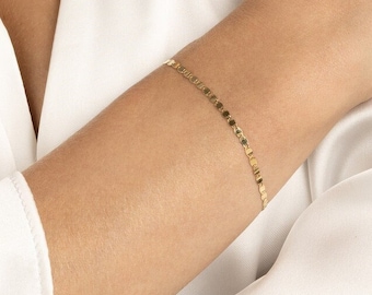 May include: A gold chain bracelet with small, square-shaped links. The bracelet is on a person's wrist.