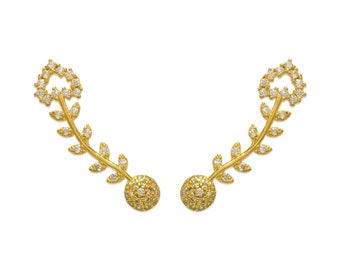 May include: A pair of gold-colored ear climbers with a leaf and floral design. The earrings feature a curved stem with small leaves and a cluster of gemstones at the base and top. The earrings are set against a white background.
