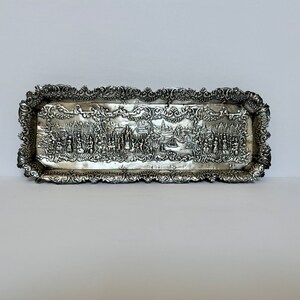 May include: A silver tray with an ornate border and a raised design of a city scene. The tray is rectangular and has a slightly curved edge.