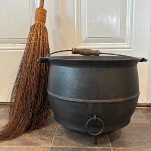 May include: A black cast iron cauldron with three legs, a metal handle featuring a wooden grip, and a circular ring attached to the front. Beside the cauldron stands a tall, long-handled broom made of brown natural straw fibers.