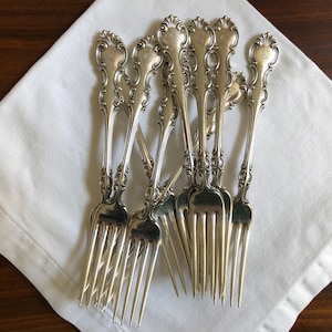 May include: A set of eight silver forks with ornate handles. The forks are arranged in a fan shape on a white napkin.