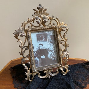 May include: Ornate gold-colored picture frame with a vintage black and white photograph. The frame has an elaborate, scrolling design. The photograph features a seated person. The frame rests on a wooden surface with a black lace cloth.
