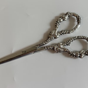May include: Silver-toned scissors with ornate handles. The handles are decorated with a grape and leaf design. The blades are straight and sharp. The scissors are likely vintage or antique, and are suitable for sewing or crafting.