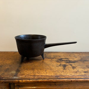 May include: A small, black cast iron pot with a long handle and three short legs. The pot has a rounded bowl shape and is likely used for cooking or heating. The pot is sitting on a wooden surface.