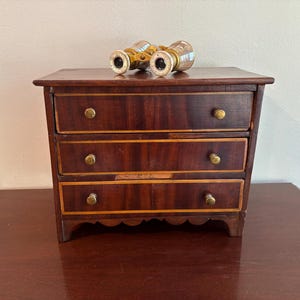 May include: A small wooden chest with three drawers, each featuring two round brass knobs and light-colored wood trim. Resting on top of the chest is a pair of vintage-style brass and mother-of-pearl opera glasses with white circular eye pieces.