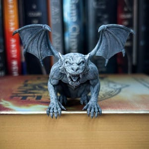 May include: A gray gargoyle figurine with outstretched wings and an open mouth, perched on a book. The detailed sculpture has a textured surface and is set against a backdrop of colorful book spines, creating a gothic aesthetic.