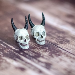 May include: Two small white skull figurines with black horns on a wooden surface.