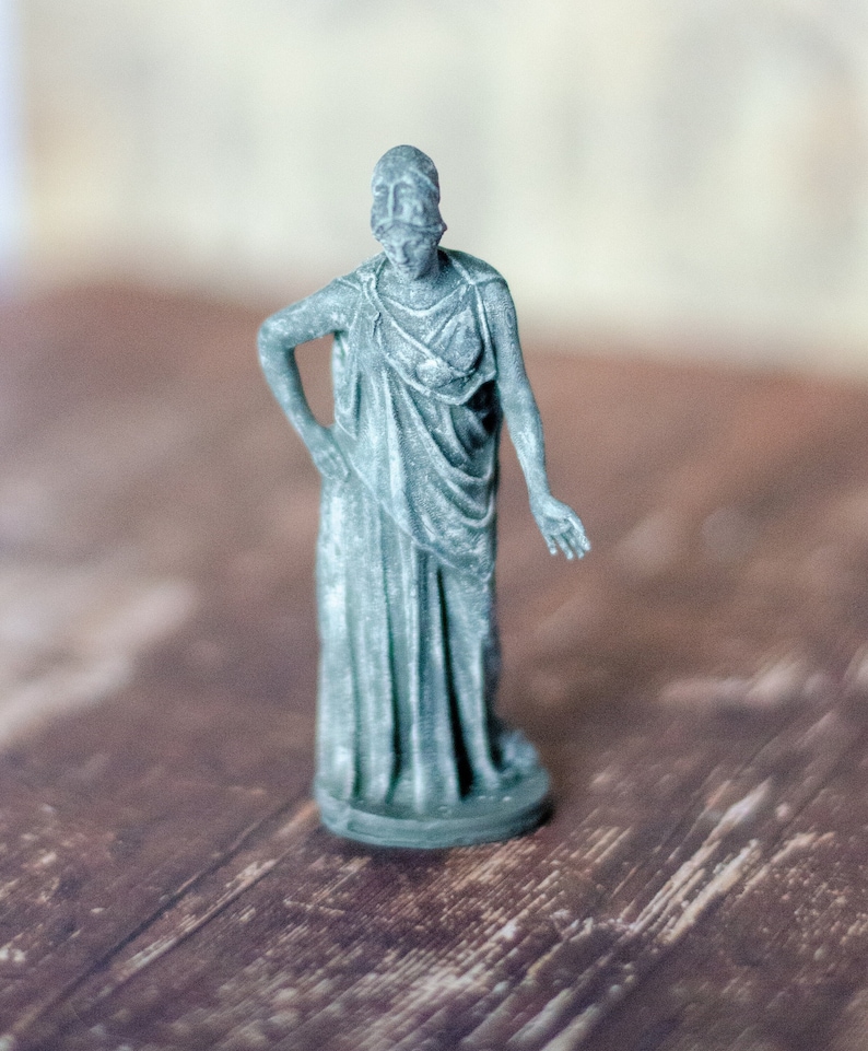 K&ouml;nnte beinhalten: Eine kleine, graue Statue einer drapierten Figur, m&ouml;glicherweise eine klassische G&ouml;ttin oder Muse. Die Figur steht mit einem Arm an der Seite und dem anderen am Ellbogen angewinkelt. Die Statue steht auf einer Holzoberfl&auml;che.